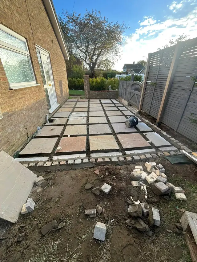 Paving slabs part-laid on a prepared base beside a bungalow, with setts stacked ready for the edge